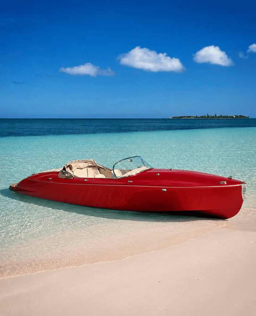 Red vintage-style boat on clear water by sandy beach and blue sky.