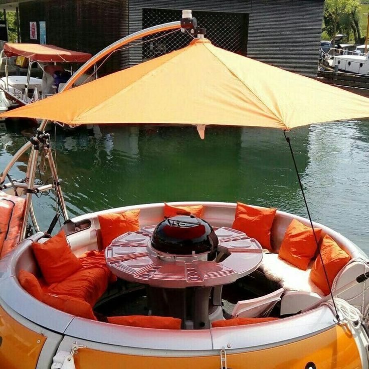Circular boat with orange umbrella, pillows, and a central grill on water.